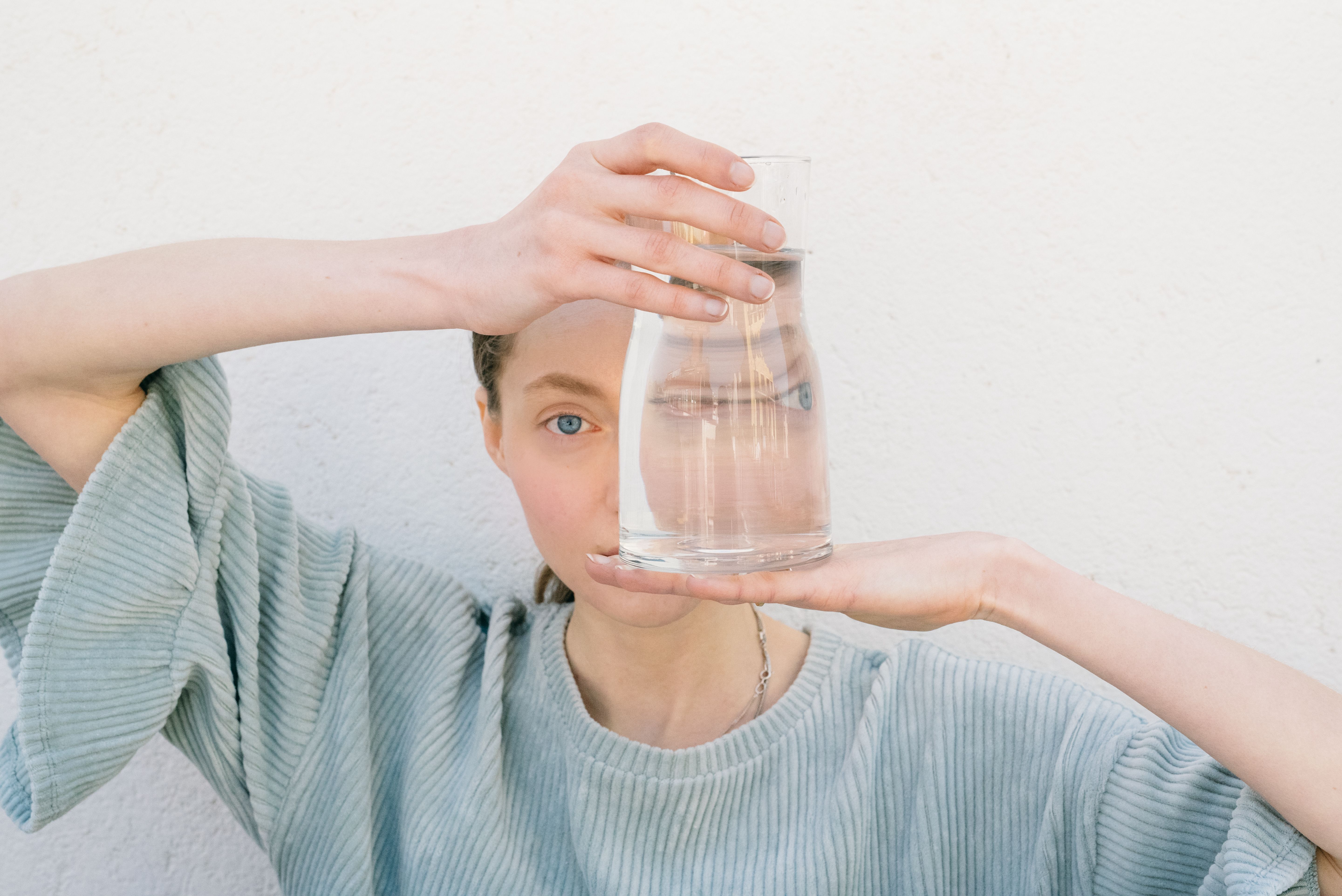 (c) pexels_leeloothefirst_Person hält ein Glas Wasser vor dem Auge. Sie trägt ein hellblaues T-Thirt mit Rüchen an den Ärmeln.