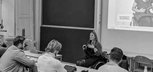 In einem Seminarraum hält eine Frau im Rollstuhl einen Vortrag vor einer Gruppe Studierender. Im Hintergrund sind eine Tafel und ein Beamer mit einer projizierten Präsentation zu sehen.