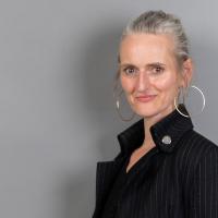 Frau mit grauen, zusammengebundenen Haaren lächelt. Sie trägt eine dunkle Jacke mit silberfarbenen Knöpfen und große goldene Creolen.