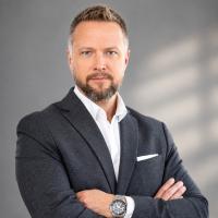 Porträt von Assoc. Prof. Mag. Dr. Michael Wininger. Er hat einen Bart und kurze, blonde Haare. Er trägt ein dunkles Sakko und ein weißes Hemd. Er blickt neutral in die Kamera. Der Hintergrund ist einfarbig in warmem Gelb.